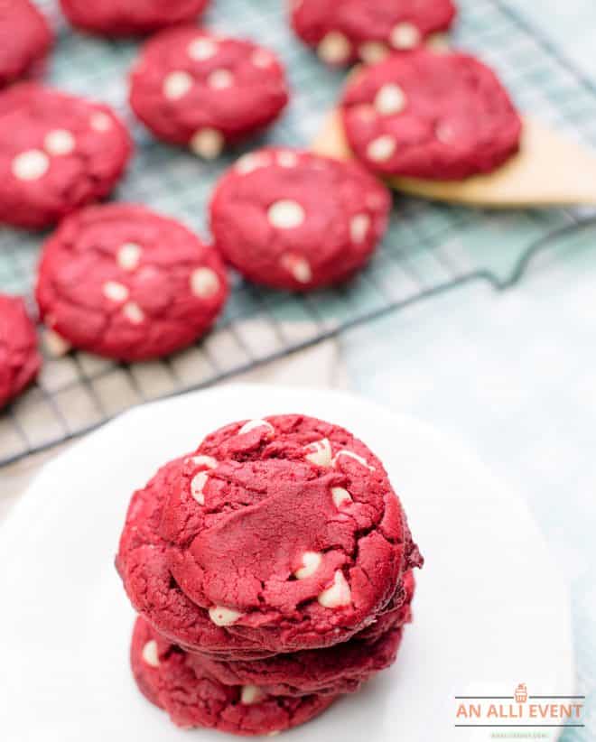 A stack of red velvet cookies on a white plate with the remaining cookies on a wire rack in the background