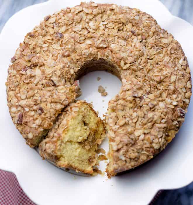 A slice of banana crunch coffee cake leaning against the rest of the cake on a white cake plate