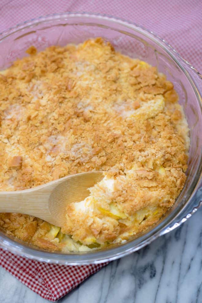Southern Squash Casserole on a red plaid dish towel with a wooden spoon lifting a serving out of the casserole dish