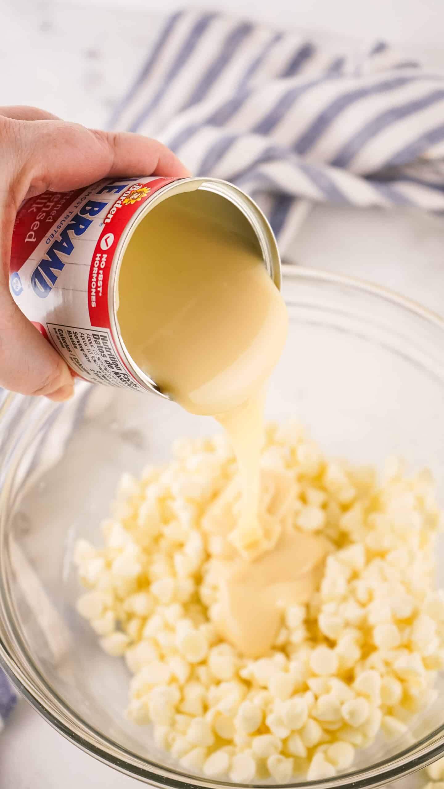 sweetened condensed milk being added to white chocolate chips in a medium glass mixing bowl