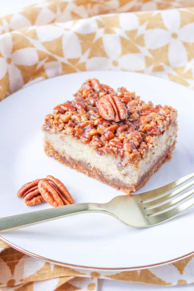 Best Pecan Pie Cheesecake Bar on a white dessert dish with a fork in front and pecans off to the side