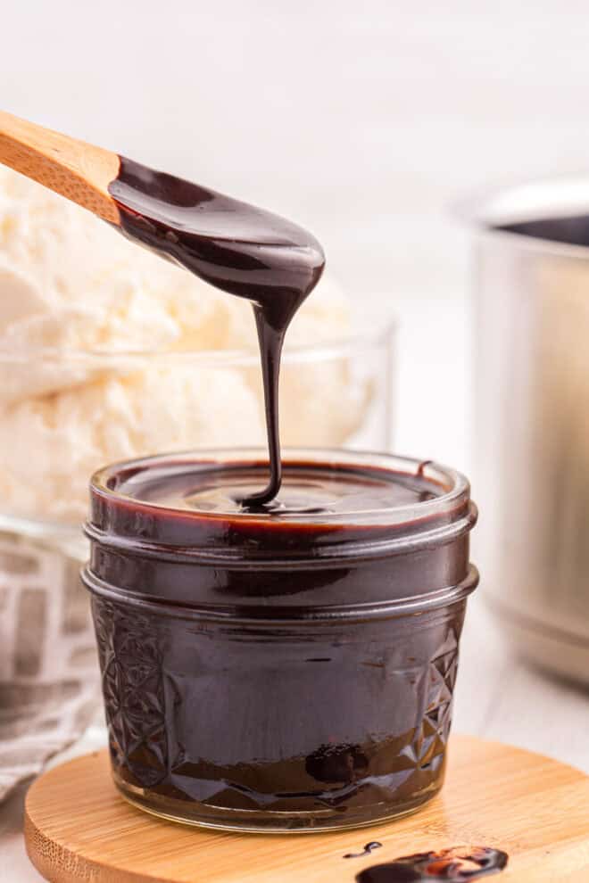 homemade hot fudge sauce in a glass jar with a wooden spoon full of sauce drizzling into the jar