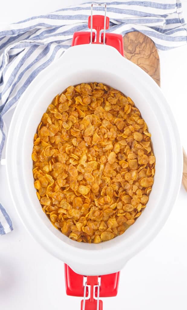 white slow cooker filled with funeral potatoes and topped with cornflakes with a blue and white dish towel to the side of the slow cooker