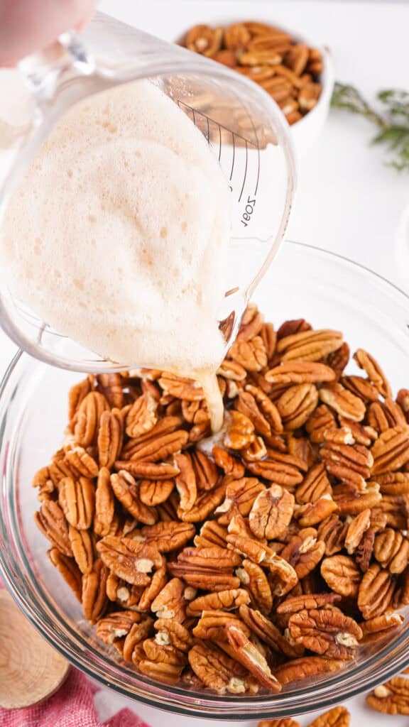 foamy egg whites being poured over raw pecans in a glass bowl