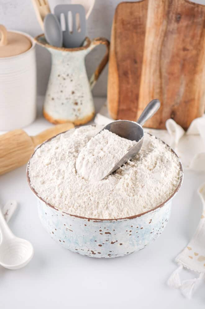 homemade self-rising flour in a rustic, white bowl with a scoop of flour on top. A cutting board and rolling pin are in the background